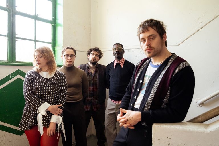 Indie rock band Wishy posing together in a stairwell in retro-style clothing near a window with green trim.